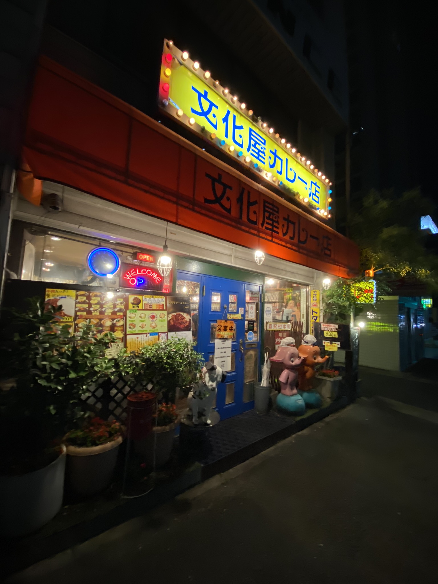 文化屋カレー店の夜の外観。黄色い看板に電球装飾、青いドア、入り口前に動物のオブジェが並んでいる