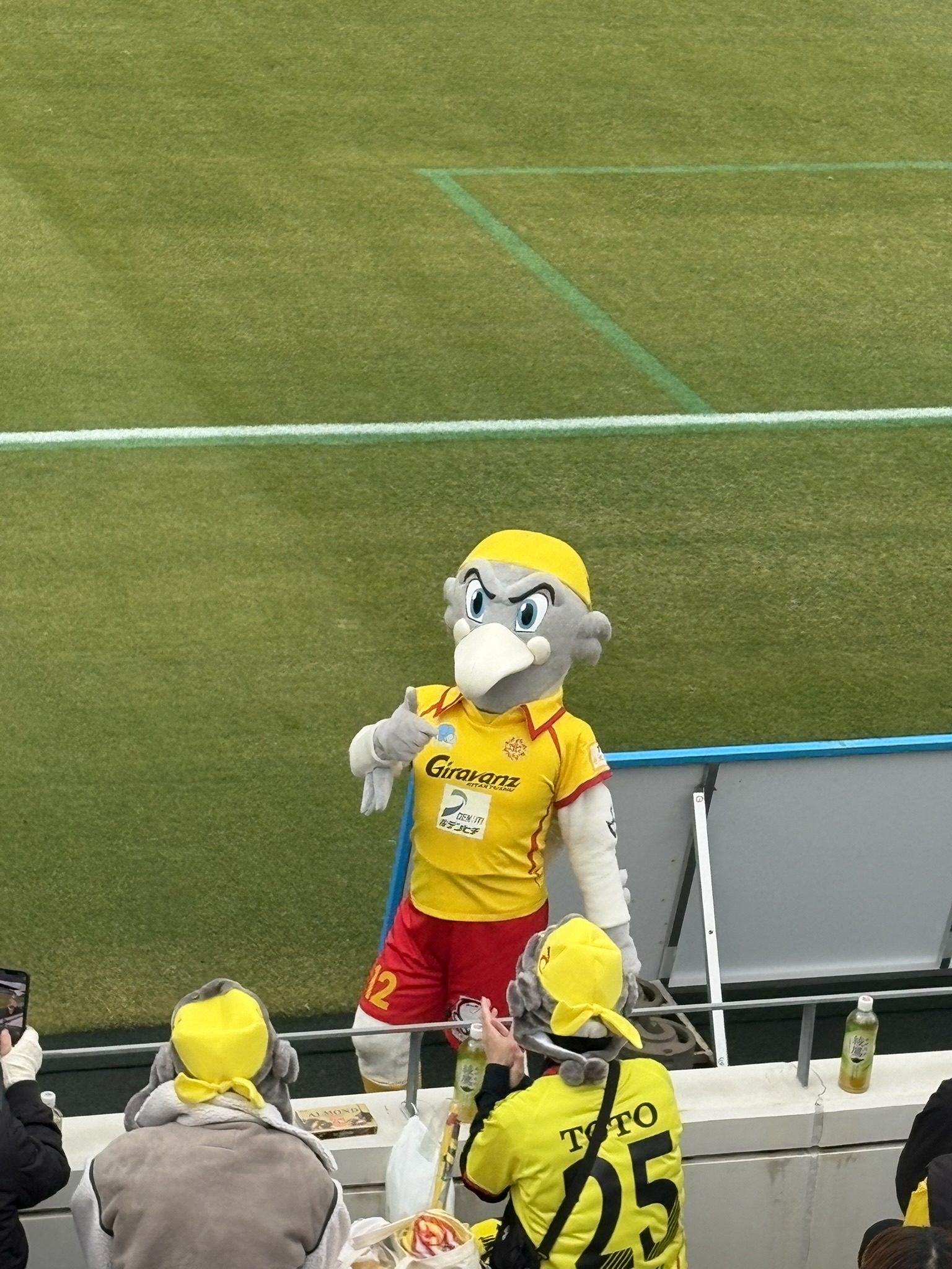 スタジアムのピッチサイドに立つ黄色いマスコットキャラクター